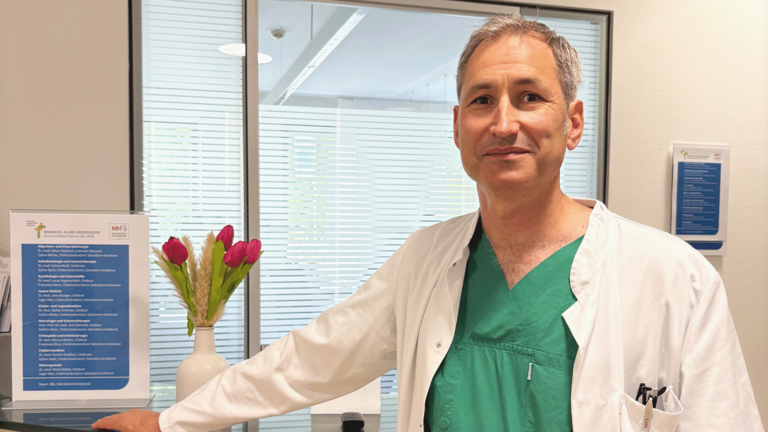 Dr. med. Oskar Rückbeil, kommissarischer Chefarzt der Allgemein- und Viszeralchirurgie, steht in Funktionskleidung am Empfangstresen in der Immanuel Klinik Rüdersdorf beim 18. Rüdersdorfer Chirurgietag zum Thema Inkontinenz und blickt in die Kamera. Dr. med. Oskar Rückbeil, kommissarischer Chefarzt der Allgemein- und Viszeralchirurgie, steht in Funktionskleidung am Empfangstresen in der Immanuel Klinik Rüdersdorf beim 18. Rüdersdorfer Chirurgietag zum Thema Inkontinenz und blickt in die Kamera.